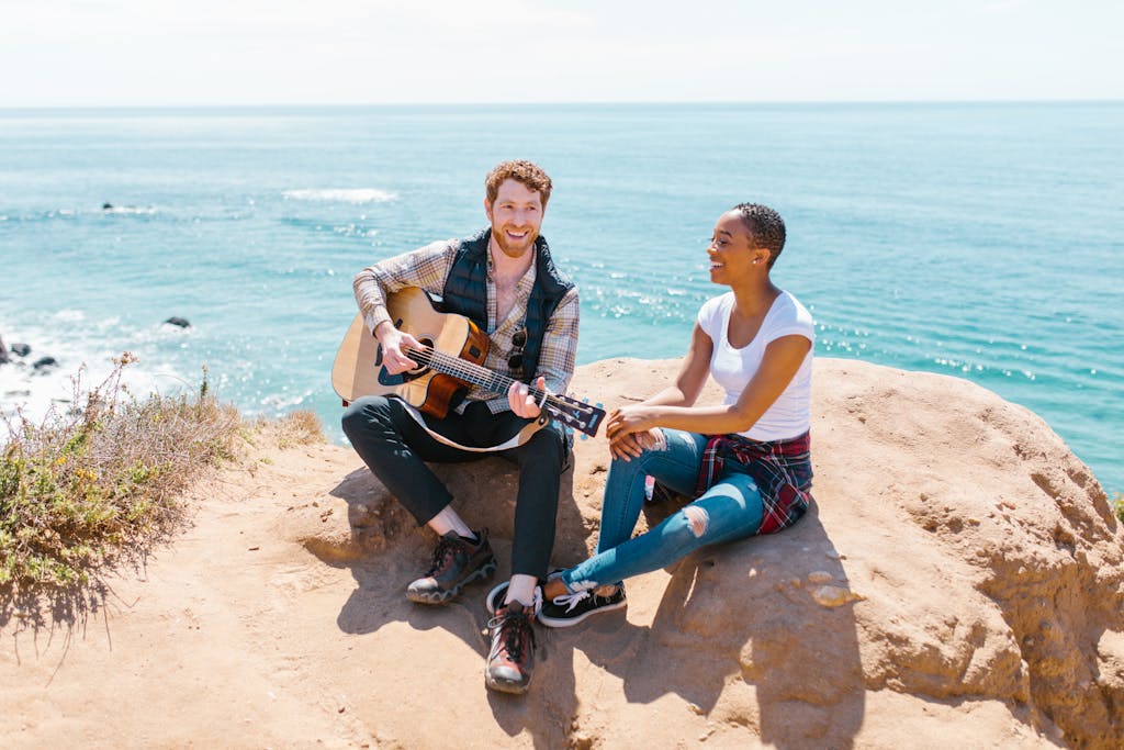 Two friends sitting on a cliff by the ocean, playing guitar and enjoying the view.